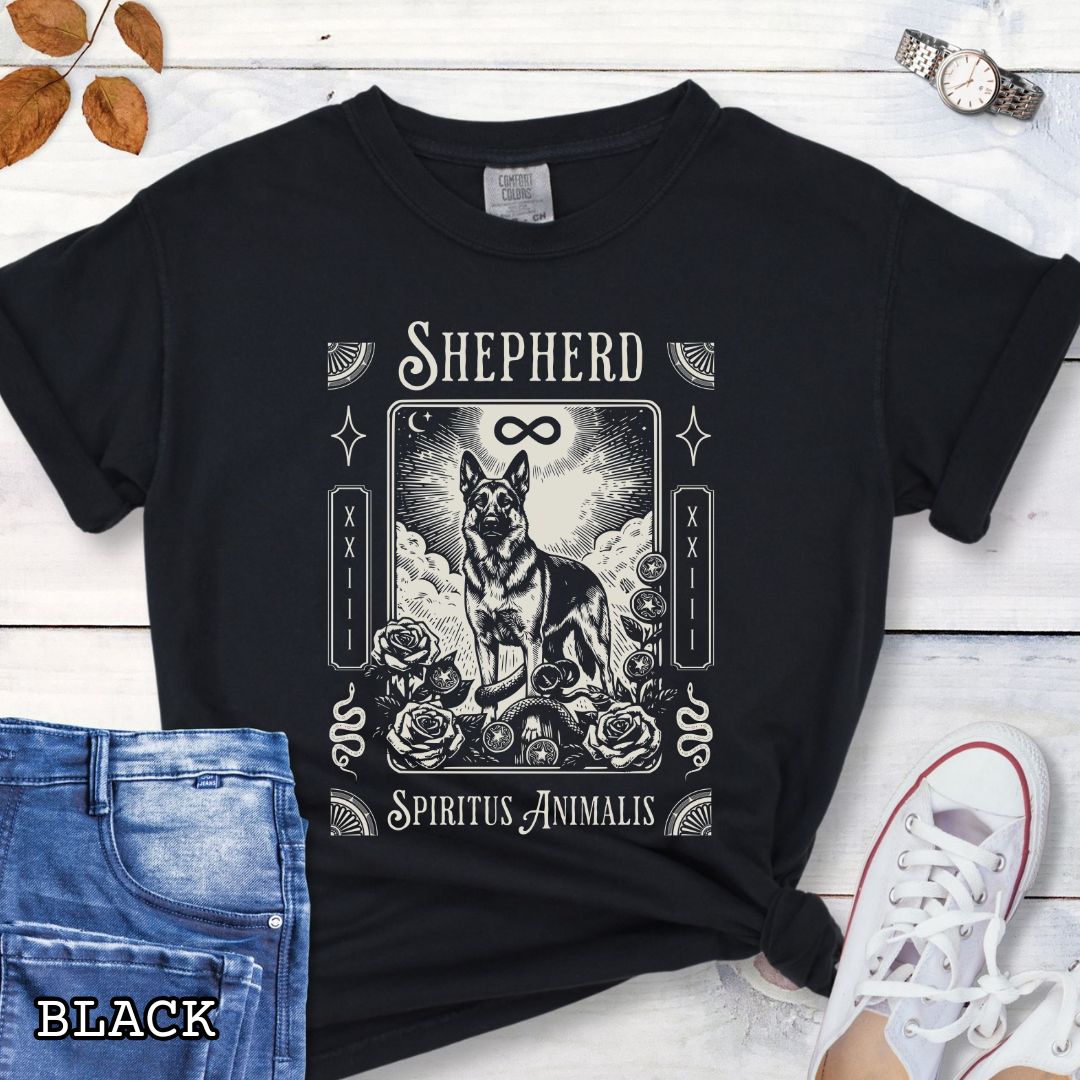 A black graphic t-shirt featuring a tarot card style illustration of a German Shepherd with the text 'SHEPHERD' and 'Spiritus Animalis'. The shirt is styled in a flat-lay arrangement on a white wood background, accompanied by blue jeans, a white sneaker, and a watch.