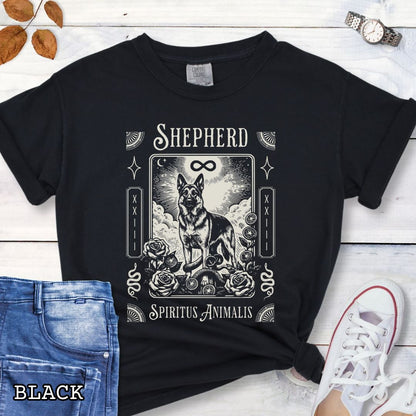 A black graphic t-shirt featuring a tarot card style illustration of a German Shepherd with the text 'SHEPHERD' and 'Spiritus Animalis'. The shirt is styled in a flat-lay arrangement on a white wood background, accompanied by blue jeans, a white sneaker, and a watch.