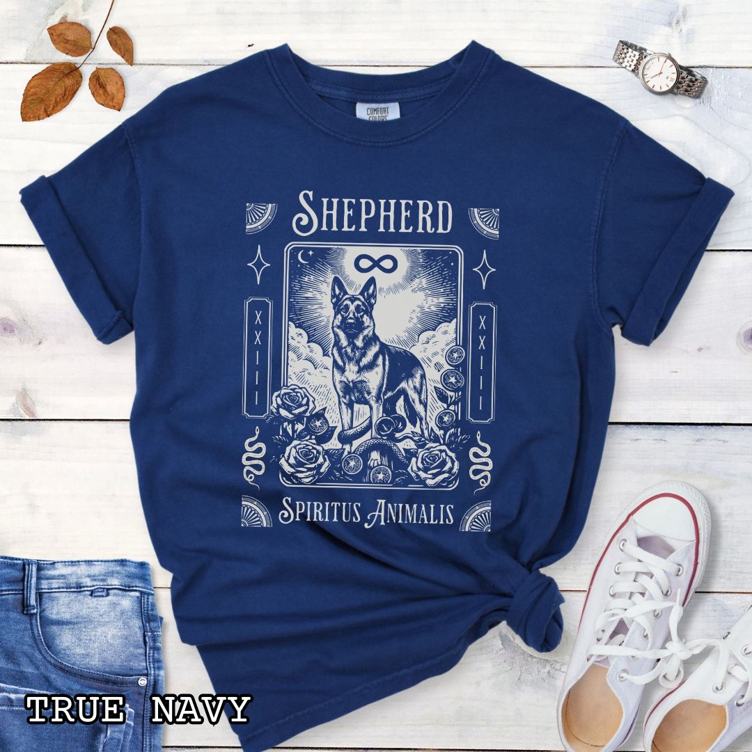A navy blue graphic t-shirt featuring a tarot card style illustration of a German Shepherd with the text 'SHEPHERD' and 'Spiritus Animalis'. The shirt is styled in a flat-lay arrangement on a white wood background, accompanied by blue jeans, a white sneaker, and a watch.