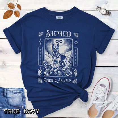 A navy blue graphic t-shirt featuring a tarot card style illustration of a German Shepherd with the text 'SHEPHERD' and 'Spiritus Animalis'. The shirt is styled in a flat-lay arrangement on a white wood background, accompanied by blue jeans, a white sneaker, and a watch.