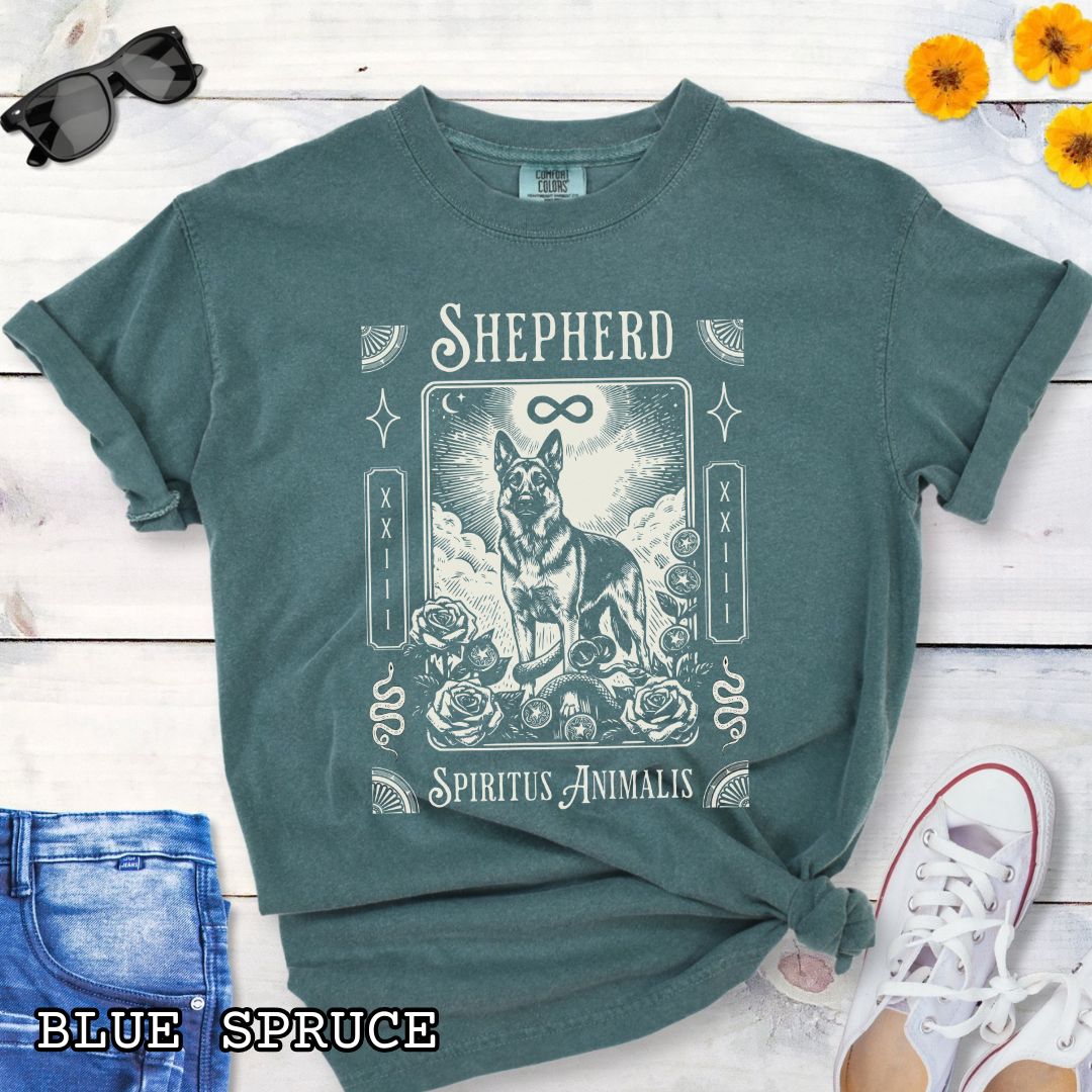 A green graphic t-shirt featuring a tarot card style illustration of a German Shepherd with the text 'SHEPHERD' and 'Spiritus Animalis'. The shirt is styled in a flat-lay arrangement on a white wood background, accompanied by blue jeans, a white sneaker, and a watch.