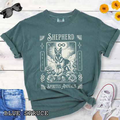 A green graphic t-shirt featuring a tarot card style illustration of a German Shepherd with the text 'SHEPHERD' and 'Spiritus Animalis'. The shirt is styled in a flat-lay arrangement on a white wood background, accompanied by blue jeans, a white sneaker, and a watch.