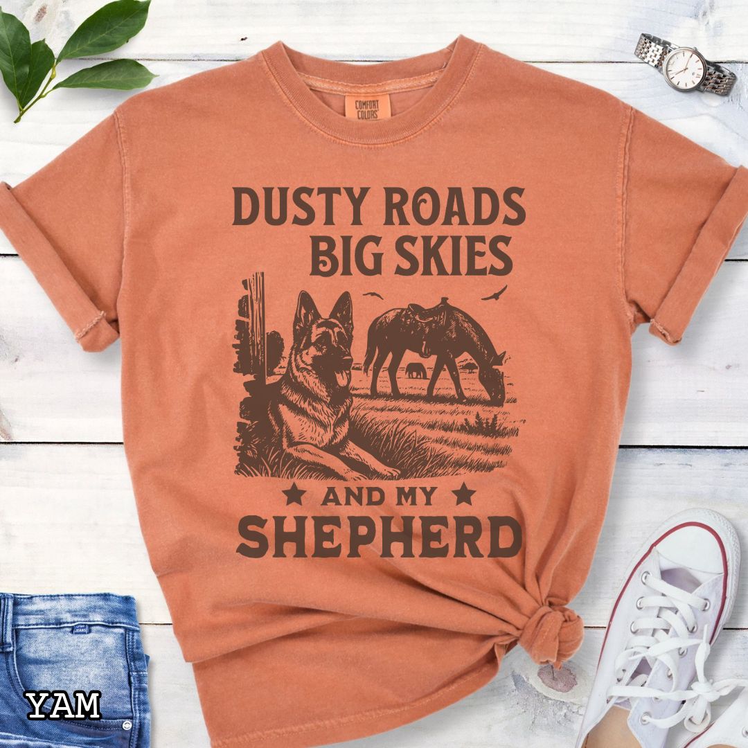 Yam-orange Comfort Colors t-shirt featuring a Western-style design with grazing horse, rustic fence, and a German Shepherd illustration; text reads “Dusty Roads, Big Skies, and My Shepherd.”