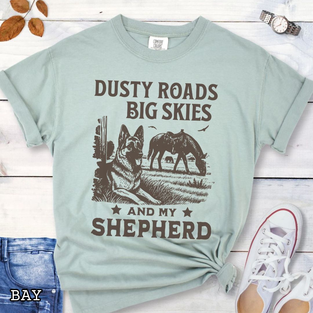 Light green Comfort Colors t-shirt featuring a Western-style design with grazing horse, rustic fence, and a German Shepherd illustration; text reads “Dusty Roads, Big Skies, and My Shepherd.”