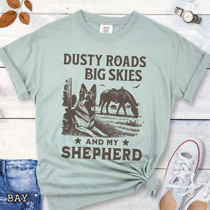 Light green Comfort Colors t-shirt featuring a Western-style design with grazing horse, rustic fence, and a German Shepherd illustration; text reads “Dusty Roads, Big Skies, and My Shepherd.”