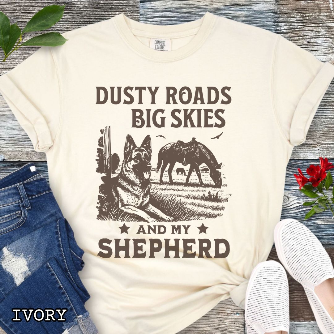 Ivory Comfort Colors t-shirt featuring a Western-style design with grazing horse, rustic fence, and a German Shepherd illustration; text reads “Dusty Roads, Big Skies, and My Shepherd.”