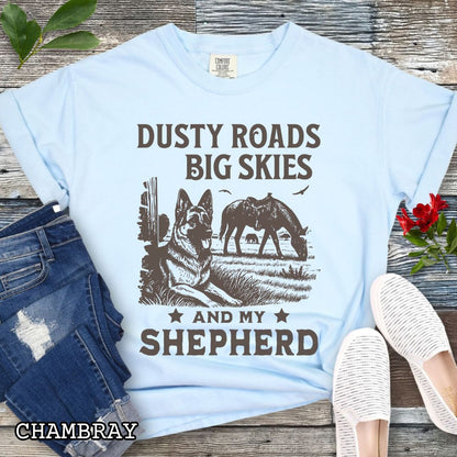 Light blue Comfort Colors t-shirt featuring a Western-style design with grazing horse, rustic fence, and a German Shepherd illustration; text reads “Dusty Roads, Big Skies, and My Shepherd.”