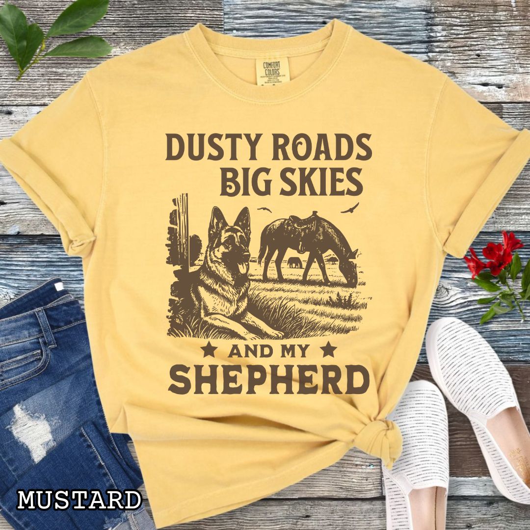 Mustard-yellow Comfort Colors t-shirt featuring a Western-style design with grazing horse, rustic fence, and a German Shepherd illustration; text reads “Dusty Roads, Big Skies, and My Shepherd.”