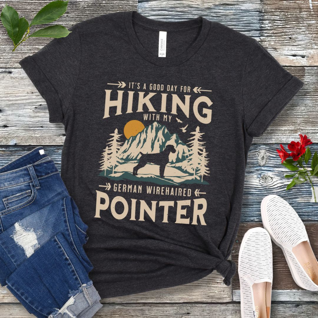A dark grey t-shirt with a vintage-style graphic print of mountains and a German Wirehaired Pointer and the text 'Hiking with my German Wirehaired Pointer'. The shirt is presented on a wooden surface with a pair of blue jeans and white shoes in the background.