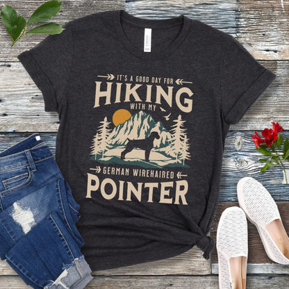 A dark grey t-shirt with a vintage-style graphic print of mountains and a German Wirehaired Pointer and the text 'Hiking with my German Wirehaired Pointer'. The shirt is presented on a wooden surface with a pair of blue jeans and white shoes in the background.