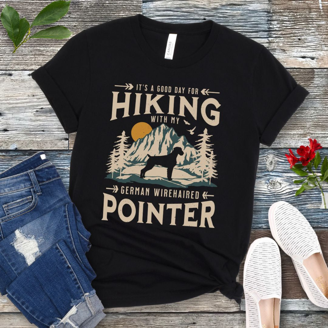 A black t-shirt with a vintage-style graphic print of mountains and a German Wirehaired Pointer and the text 'Hiking with my German Wirehaired Pointer'. The shirt is presented on a wooden surface with a pair of blue jeans and white shoes in the background.