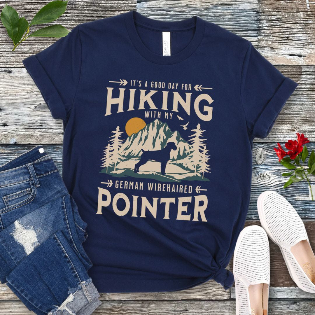 A navy t-shirt with a vintage-style graphic print of mountains and a German Wirehaired Pointer and the text 'Hiking with my German Wirehaired Pointer'. The shirt is presented on a wooden surface with a pair of blue jeans and white shoes in the background.
