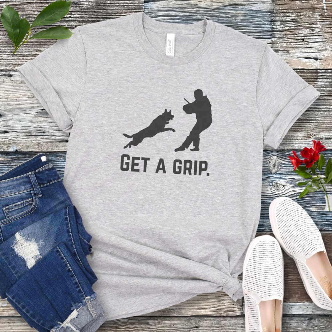 Gray t-shirt with bite work graphic of a protection dog and K9 helper and the text 'GET A GRIP.' on a wooden surface with jeans and shoes.