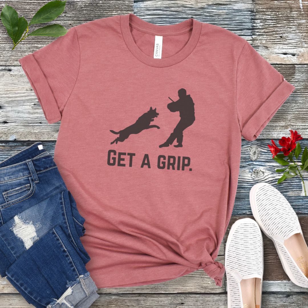 Mauve t-shirt with bite work graphic of a protection dog and K9 helper and the text 'GET A GRIP.' on a wooden surface with jeans and shoes.