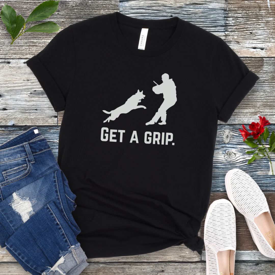 Black t-shirt with bite work graphic of a protection dog and K9 helper and the text 'GET A GRIP.' on a wooden surface with jeans and shoes.