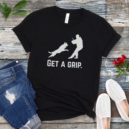 Black t-shirt with bite work graphic of a protection dog and K9 helper and the text 'GET A GRIP.' on a wooden surface with jeans and shoes.