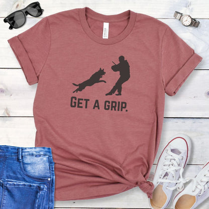 Mauve t-shirt with bite work graphic of a protection dog and K9 helper and the text 'GET A GRIP.' on a wooden surface with jeans and shoes.