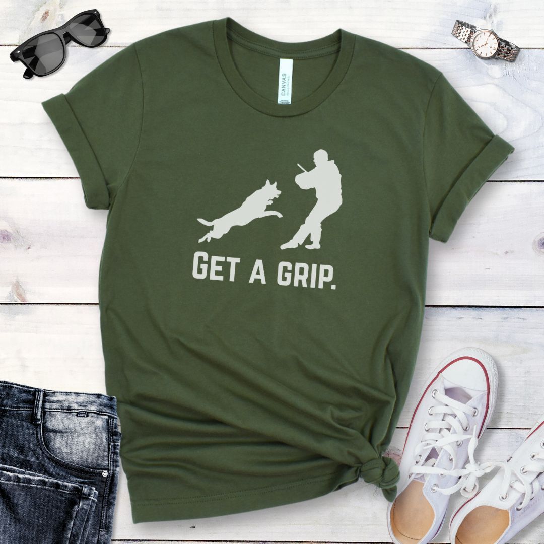 Military green t-shirt with bite work graphic of a protection dog and K9 helper and the text 'GET A GRIP.' on a wooden surface with jeans and shoes.