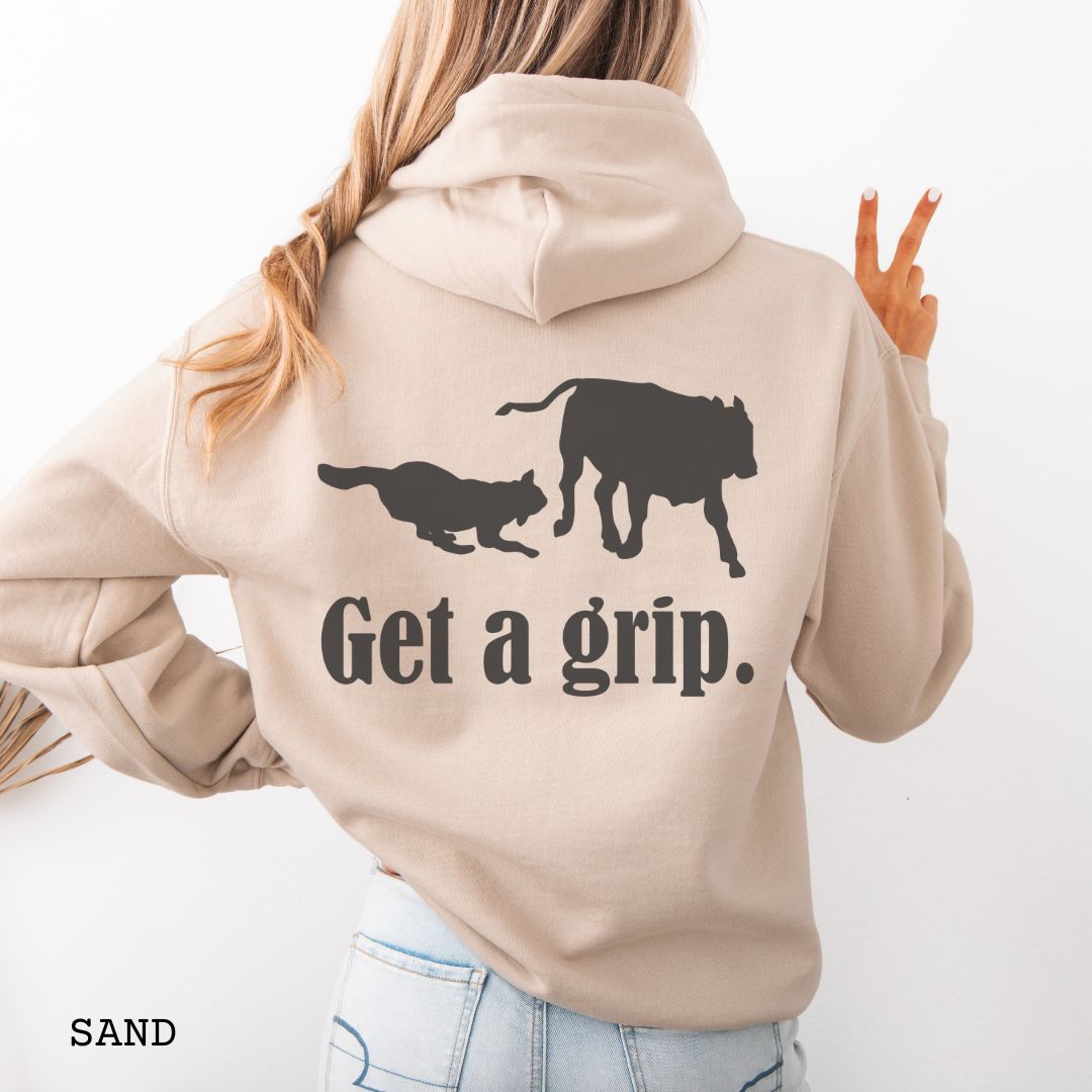 Beige back print hoodie with a graphic of a Heeler dog herding a cow and the text 'Get a grip.' Worn by a person.