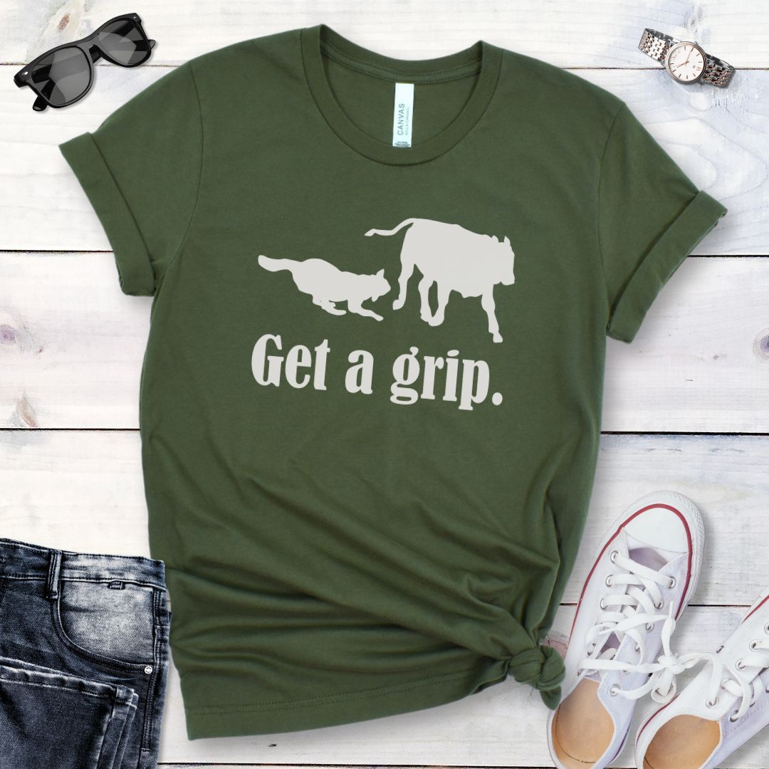A military green t-shirt with a graphic of a dog herding a cow and the humorous text 'Get a grip.' The t-shirt is presented in a casual style with rolled-up sleeves, accompanied by jeans and white shoes.