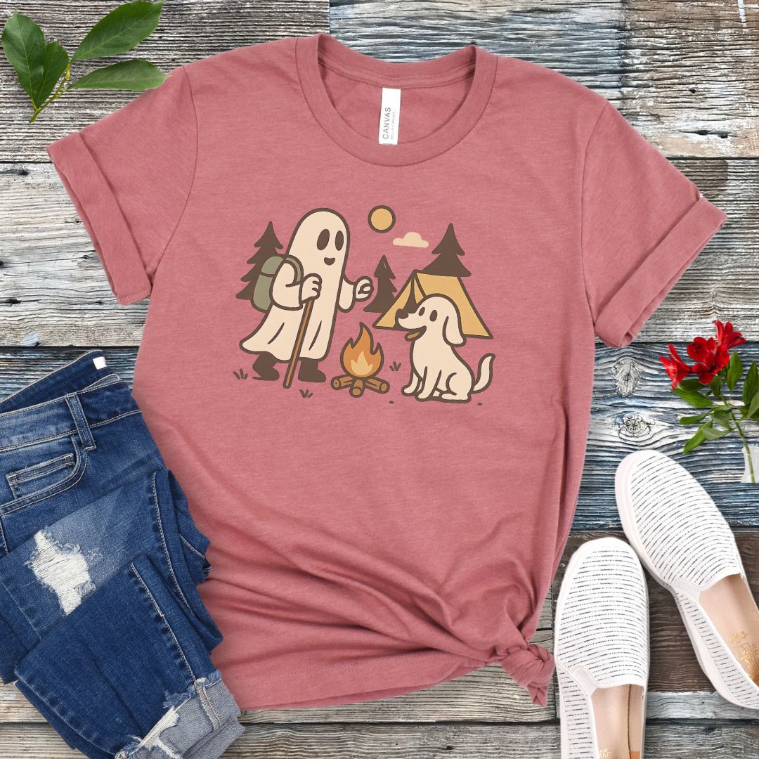 A mauve t-shirt with a graphic of a ghost roasting a marshmallow over a fire, accompanied by a dog, in a camping setting. The t-shirt is presented flat with a pair of jeans and white shoes.