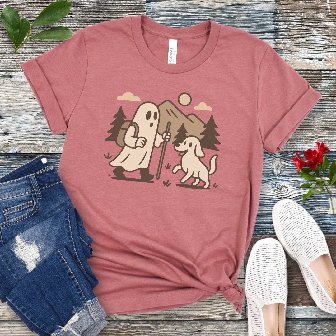 A mauve t-shirt with a graphic print of a ghost and a dog hiking in the woods, displayed with a pair of blue jeans and white shoes.