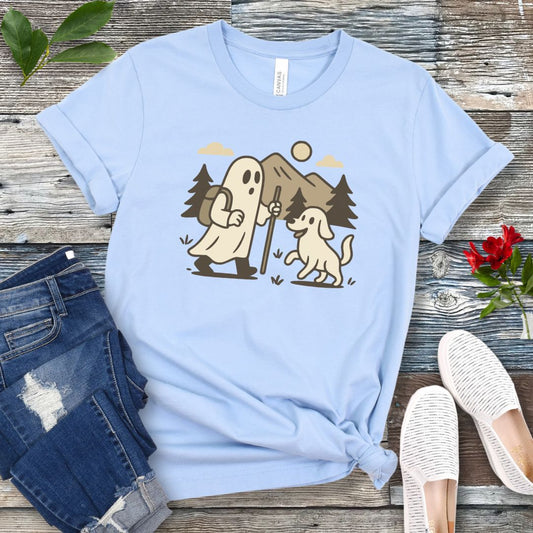 A light blue t-shirt with a graphic print of a ghost and a dog hiking in the woods, displayed with a pair of blue jeans and white shoes.