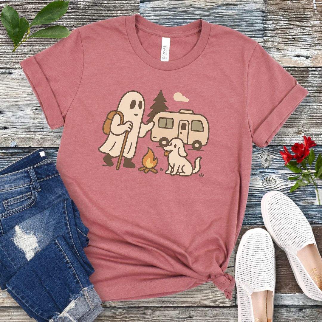 A mauve T-shirt with a graphic design featuring a ghost, a dog, and a recreational vehicle (RV) with a campfire in front of it, presented on a wooden surface with jeans and white shoes in the background.