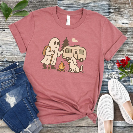 A mauve T-shirt with a graphic design featuring a ghost, a dog, and a recreational vehicle (RV) with a campfire in front of it, presented on a wooden surface with jeans and white shoes in the background.