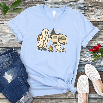 A light blue T-shirt with a graphic design featuring a ghost, a dog, and a recreational vehicle (RV) with a campfire in front of it, presented on a wooden surface with jeans and white shoes in the background.