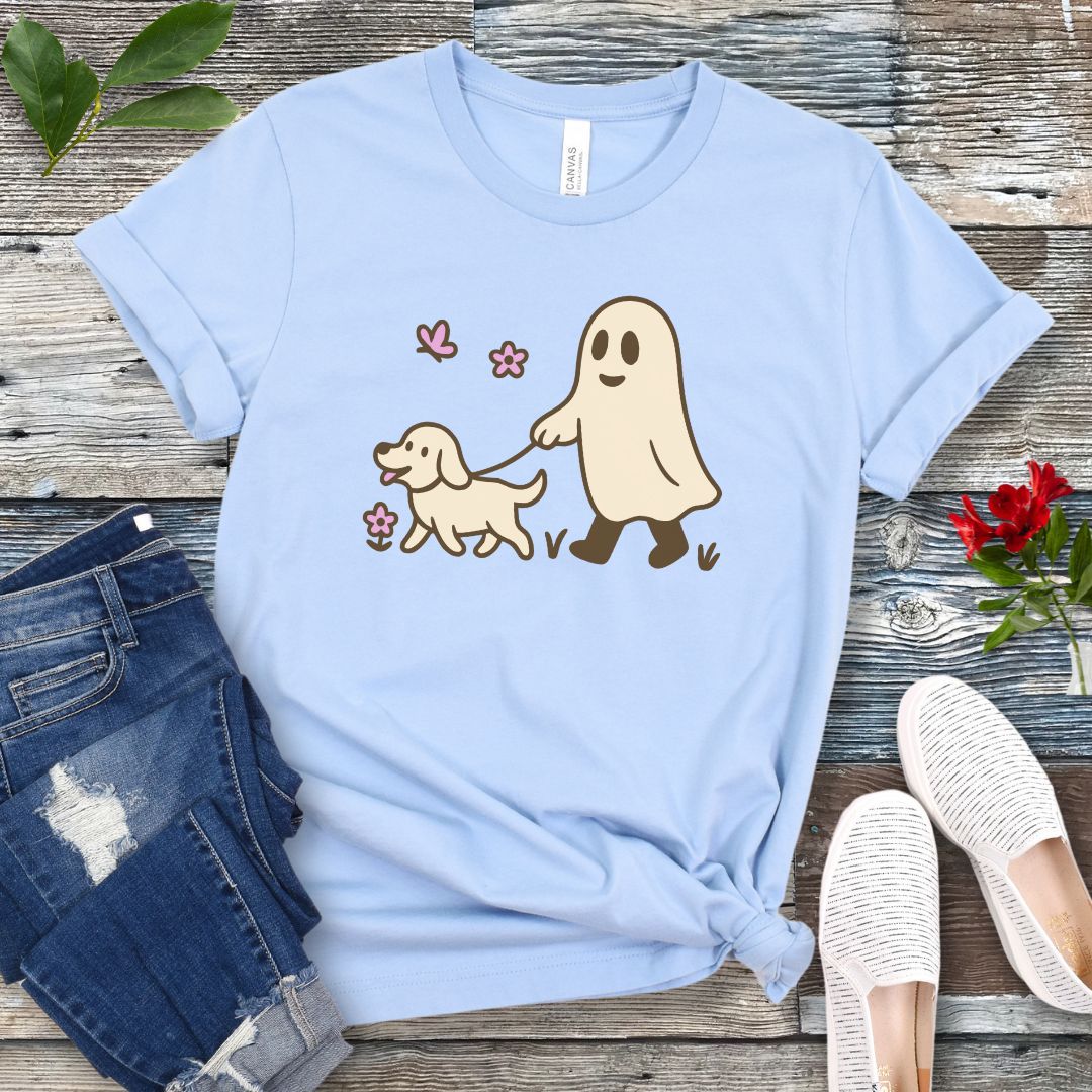 A light blue dog lover t-shirt with a graphic of a ghost walking a dog, accompanied by two pink flowers, displayed on a wooden surface with white sneakers and rolled-up blue jeans in the background.