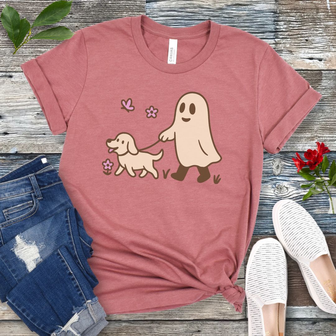A mauve dog lover t-shirt with a graphic of a ghost walking a dog, accompanied by two pink flowers, displayed on a wooden surface with white sneakers and rolled-up blue jeans in the background.