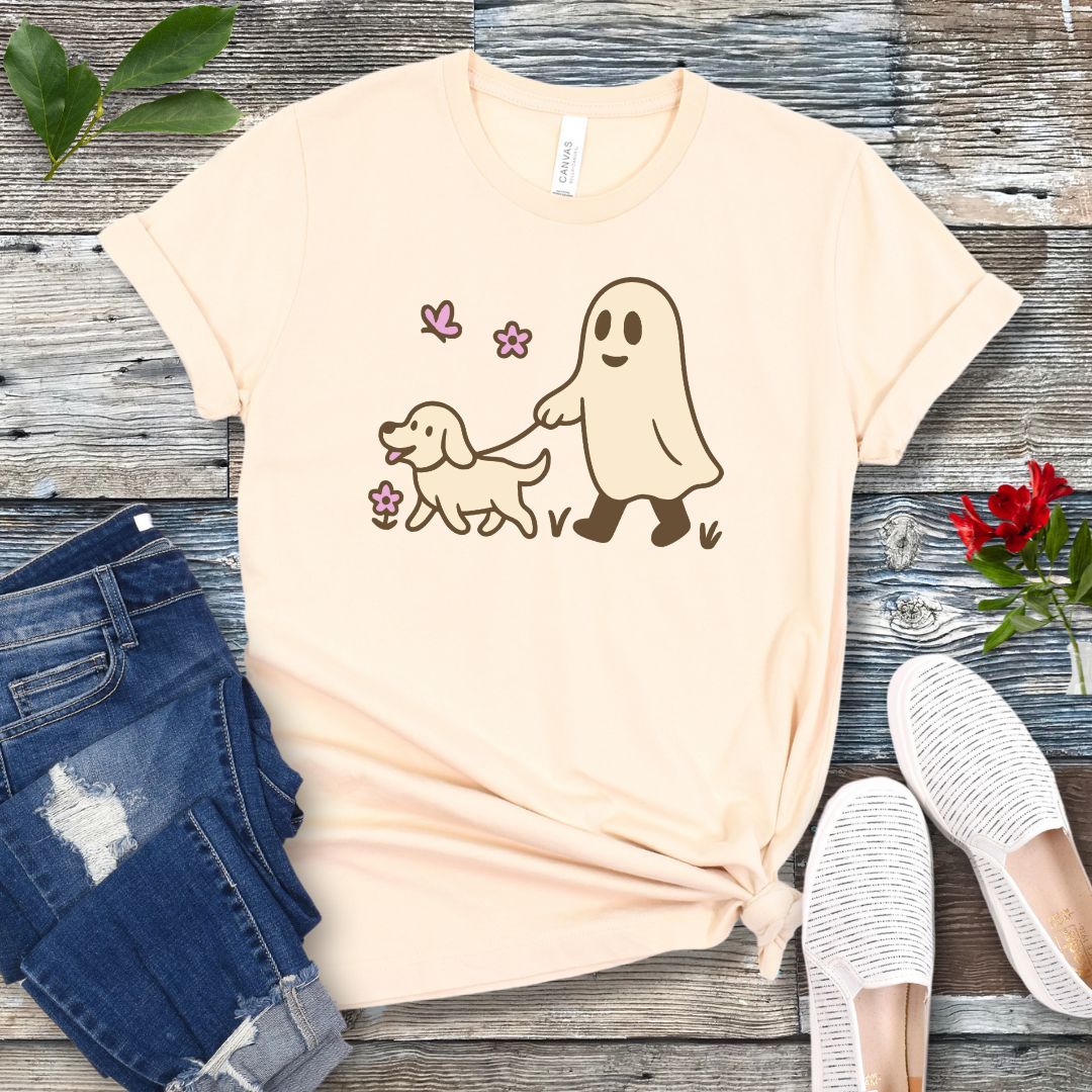 A beige dog lover t-shirt with a graphic of a ghost walking a dog, accompanied by two pink flowers, displayed on a wooden surface with white sneakers and rolled-up blue jeans in the background.