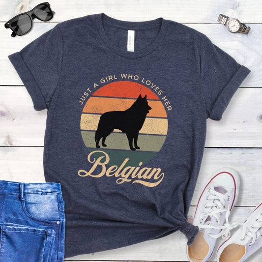 Heather navy blue t-shirt for Belgian Sheepdog lovers with vintage sunset graphic and Belgian Sheepdog silhouette, and text 'Just a Girl Who Loves Her Belgian', shown on a wooden surface with accessories.