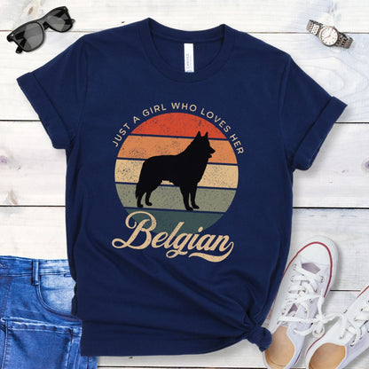 Navy blue t-shirt for Belgian Sheepdog lovers with vintage sunset graphic and Belgian Sheepdog silhouette, and text 'Just a Girl Who Loves Her Belgian', shown on a wooden surface with accessories.
