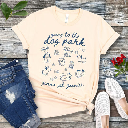 A beige t-shirt for dog lovers with 'Going to the dog park, gonna get zoomies' text in playful handwritten font and hand-drawn doodles related to dogs, laid out flat with a pair of blue jeans and white shoes in the background.
