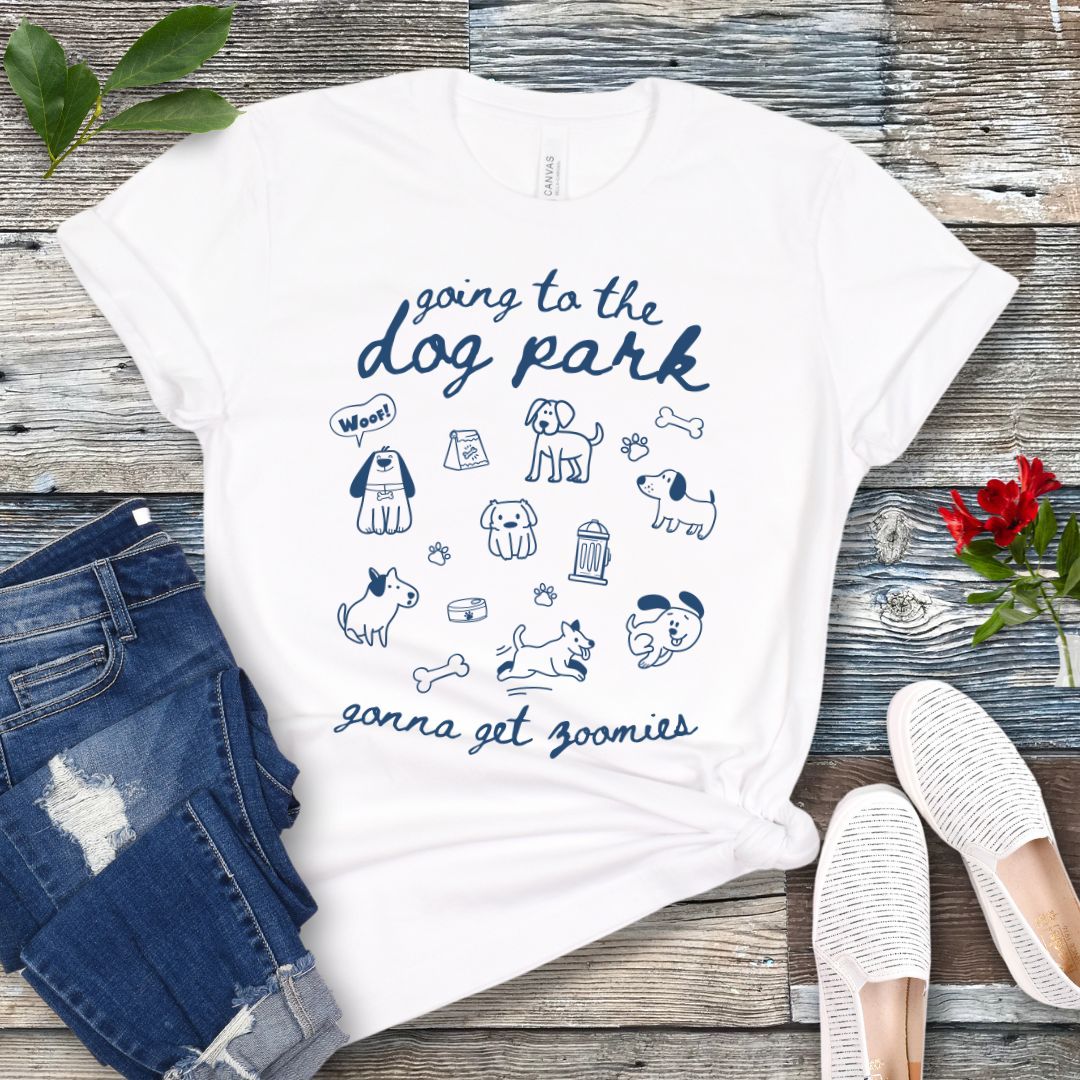 A white t-shirt for dog lovers with 'Going to the dog park, gonna get zoomies' text in playful handwritten font and hand-drawn doodles related to dogs, laid out flat with a pair of blue jeans and white shoes in the background.