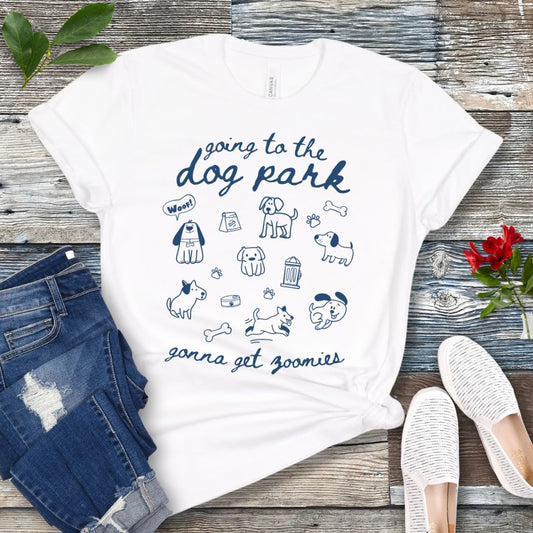 A white t-shirt for dog lovers with 'Going to the dog park, gonna get zoomies' text in playful handwritten font and hand-drawn doodles related to dogs, laid out flat with a pair of blue jeans and white shoes in the background.