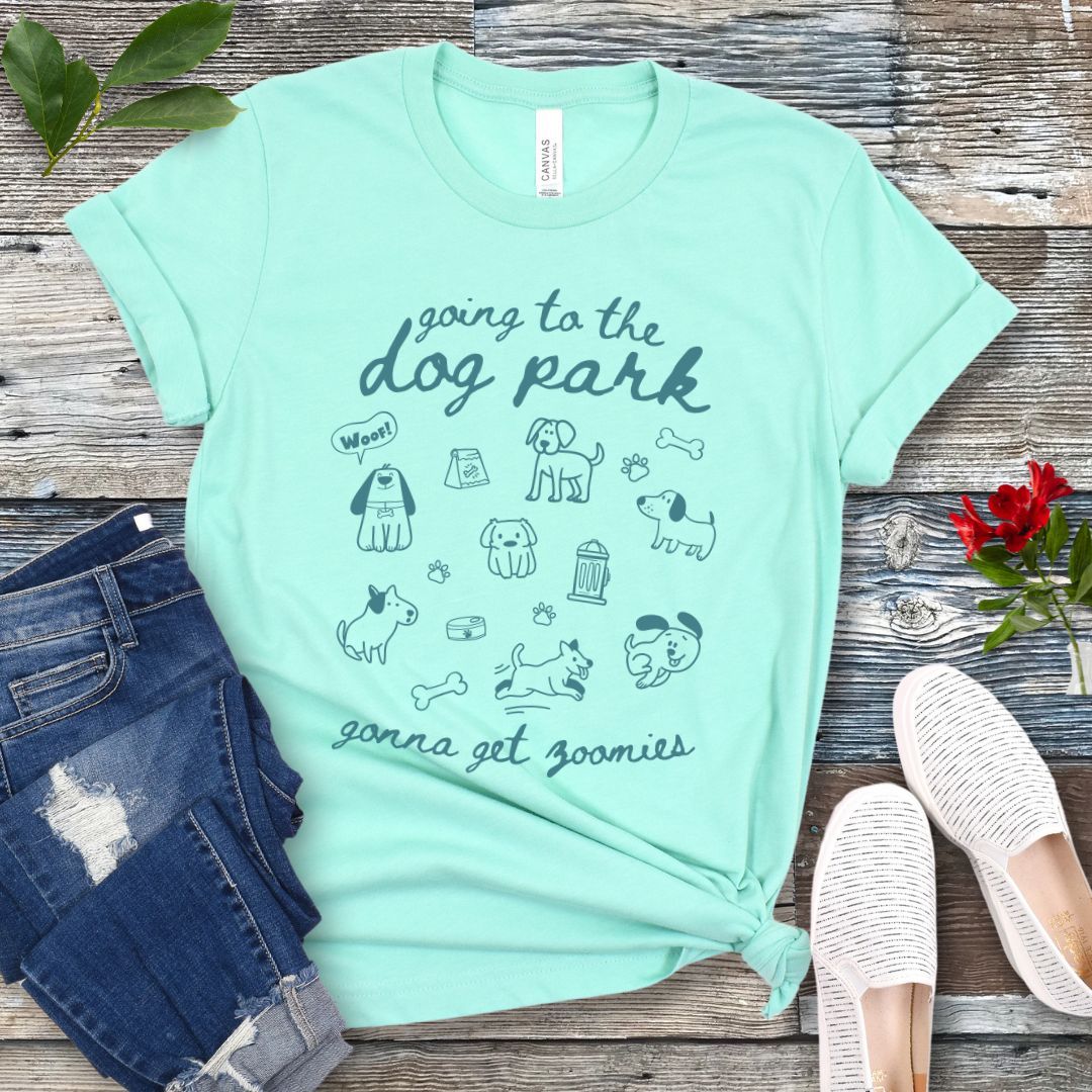 A mint green t-shirt for dog lovers with 'Going to the dog park, gonna get zoomies' text in playful handwritten font and hand-drawn doodles related to dogs, laid out flat with a pair of blue jeans and white shoes in the background.