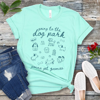 A mint green t-shirt for dog lovers with 'Going to the dog park, gonna get zoomies' text in playful handwritten font and hand-drawn doodles related to dogs, laid out flat with a pair of blue jeans and white shoes in the background.