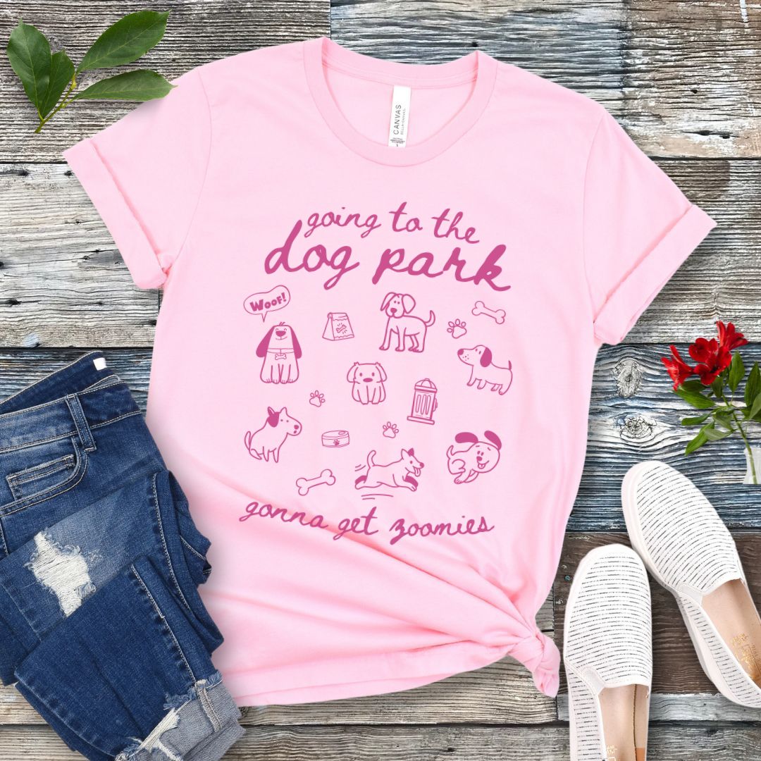 A pink t-shirt for dog lovers with 'Going to the dog park, gonna get zoomies' text in playful handwritten font and hand-drawn doodles related to dogs, laid out flat with a pair of blue jeans and white shoes in the background.