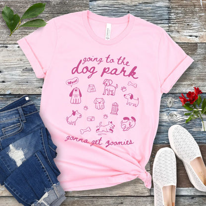 A pink t-shirt for dog lovers with 'Going to the dog park, gonna get zoomies' text in playful handwritten font and hand-drawn doodles related to dogs, laid out flat with a pair of blue jeans and white shoes in the background.