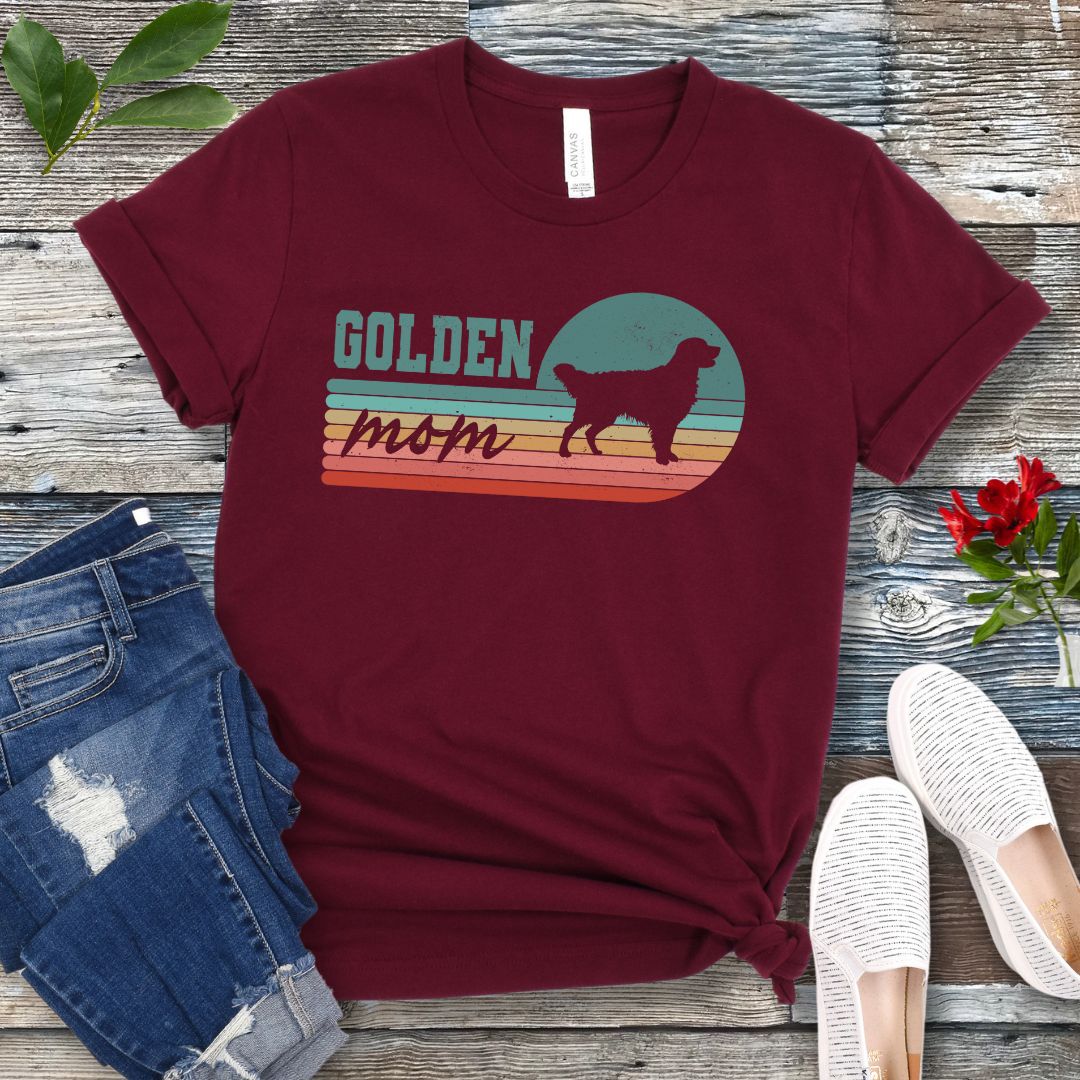 Maroon t-shirt for Golden Retriever dog lovers with 'Golden Mom' text over retro sunset stripes design on a wooden surface with jeans and shoes.