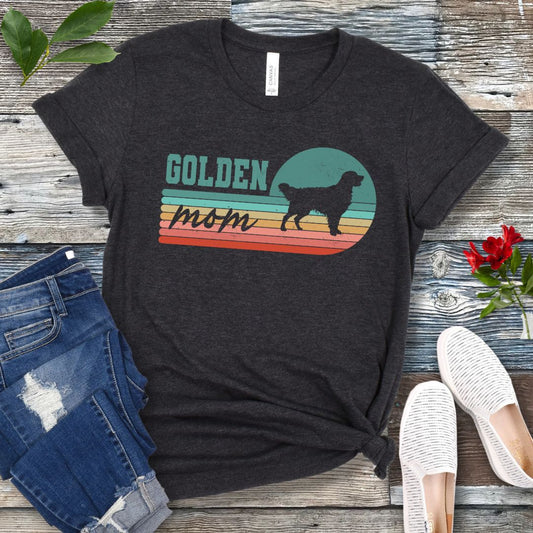 Dark grey t-shirt for Golden Retriever dog lovers with 'Golden Mom' text over retro sunset stripes design on a wooden surface with jeans and shoes.
