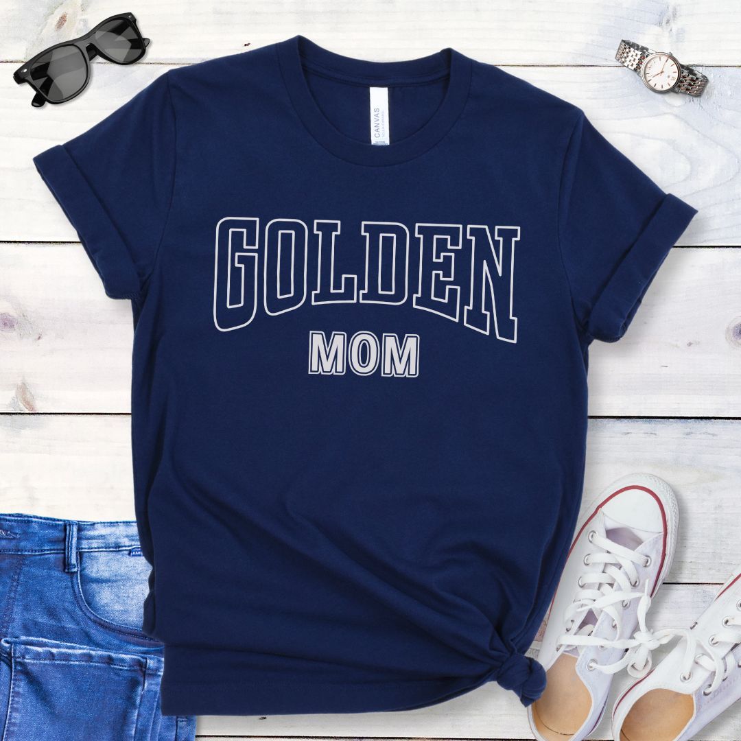 Navy blue t-shirt for Golden Retriever lovers with 'GOLDEN MOM' text in varsity style lettering, shown on a wooden surface with jeans, sneakers, and sunglasses.