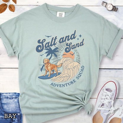 Light green Comfort Colors t-shirt for Golden Retriever lovers with beach-themed graphic of a surfing Golden, waves and sunset, and the text 'Salt and Sand Adventure Squad' in retro lettering, shown on a wooden surface with accessories.