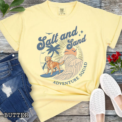 Yellow Comfort Colors t-shirt for Golden Retriever lovers with beach-themed graphic of a surfing Golden, waves and sunset, and the text 'Salt and Sand Adventure Squad' in retro lettering, shown on a wooden surface with accessories.