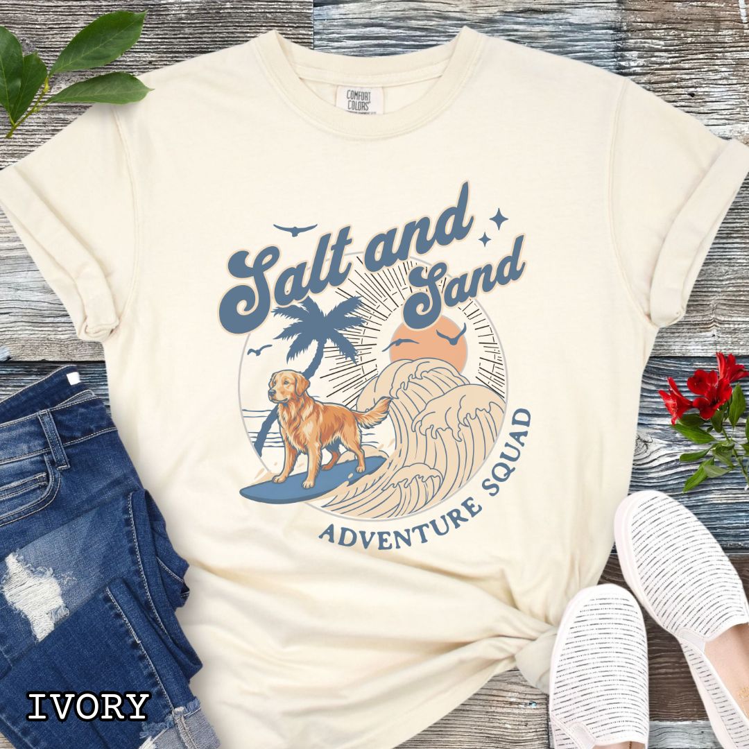 Beige Comfort Colors t-shirt for Golden Retriever lovers with beach-themed graphic of a surfing Golden, waves and sunset, and the text 'Salt and Sand Adventure Squad' in retro lettering, shown on a wooden surface with accessories.