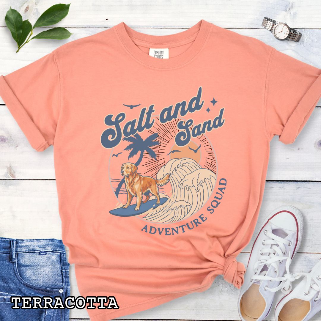 Orange Comfort Colors t-shirt for Golden Retriever lovers with beach-themed graphic of a surfing Golden, waves and sunset, and the text 'Salt and Sand Adventure Squad' in retro lettering, shown on a wooden surface with accessories.