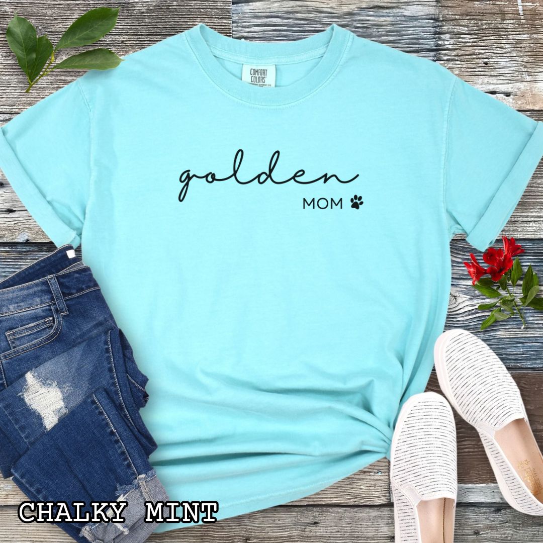 Mint Comfort Colors unisex t-shirt for Golden Retriever lovers with 'golden mom' text in feminine script and a small paw print, shown on a wooden surface with accessories. Perfect dog owner gift for Golden dog moms.